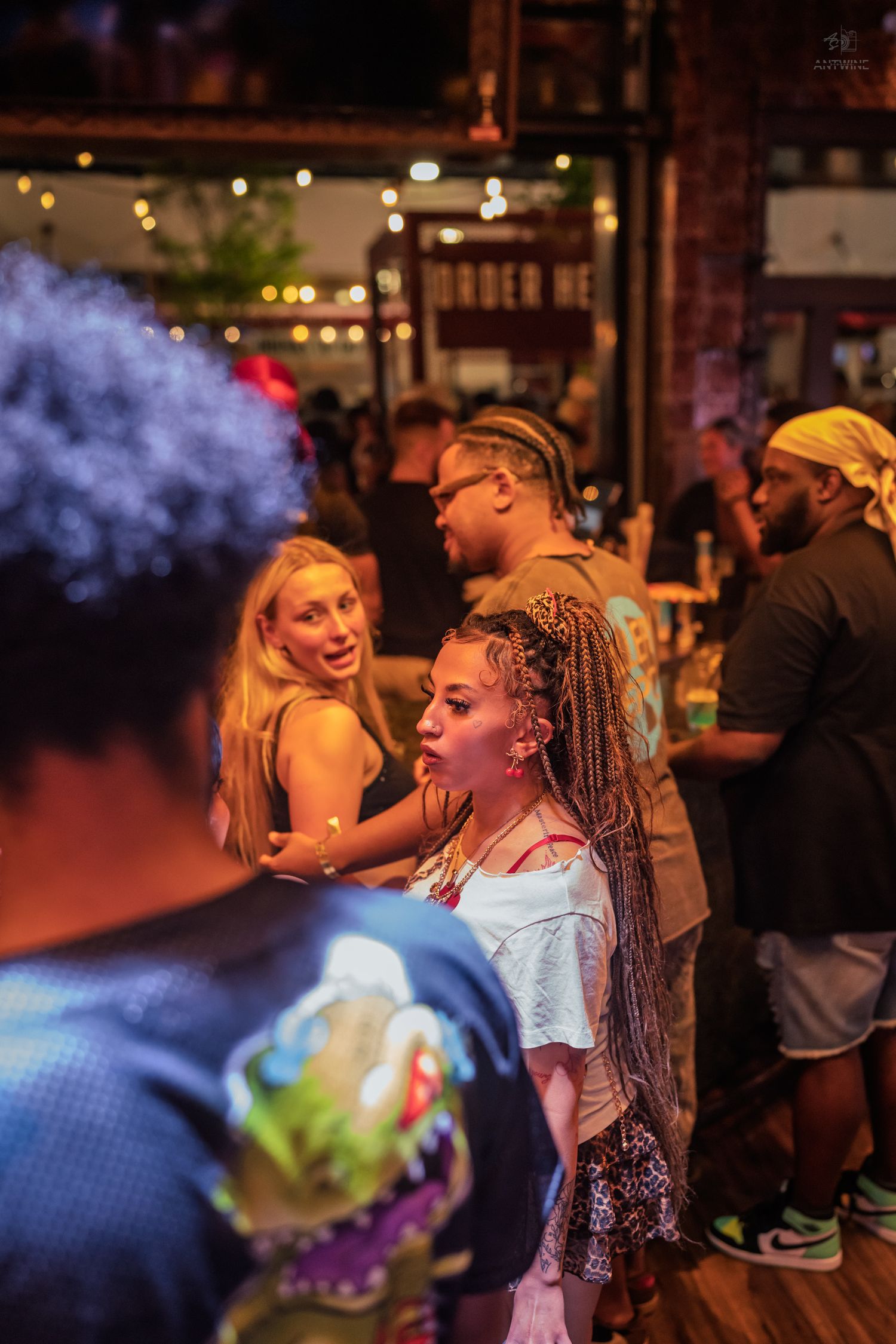 Women with colorful braids and blonde hair socialize at a lively outdoor bar event under warm string lights.