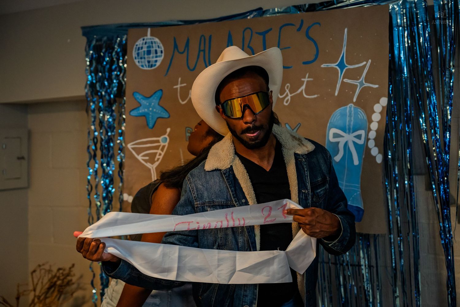Man in cowboy hat and sunglasses examining a birthday sash at a blue-themed party.