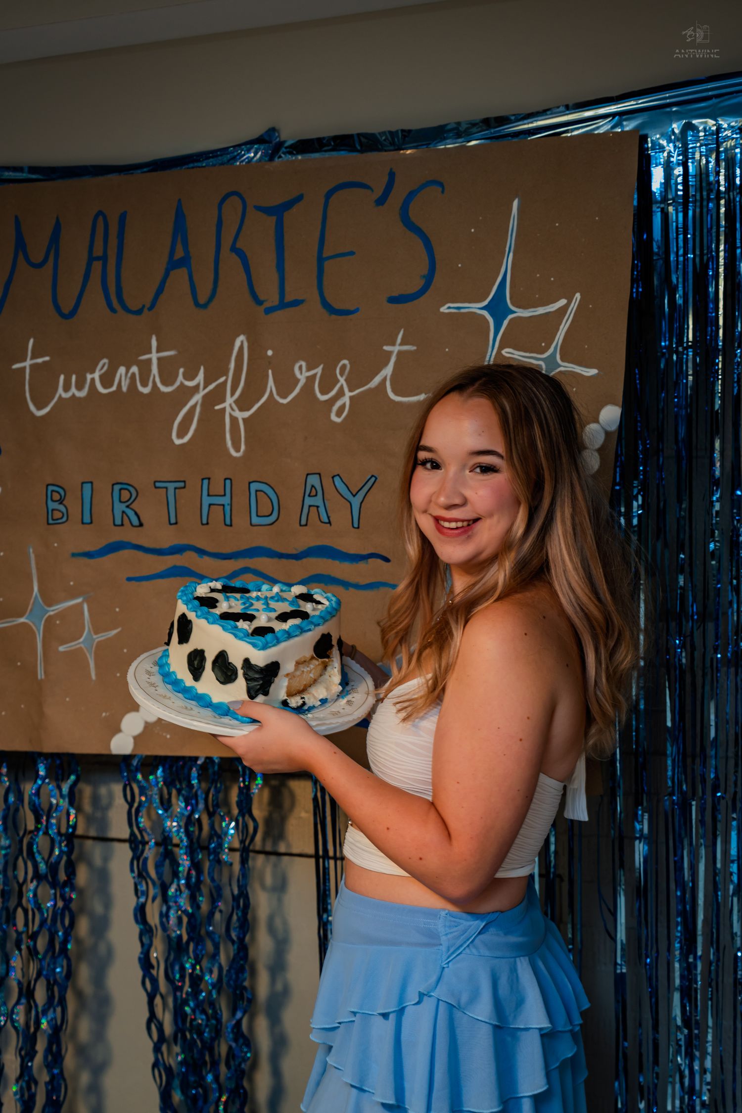 Smiling young woman holds heart-shaped cake at Malarie's 21st birthday party with blue backdrop.