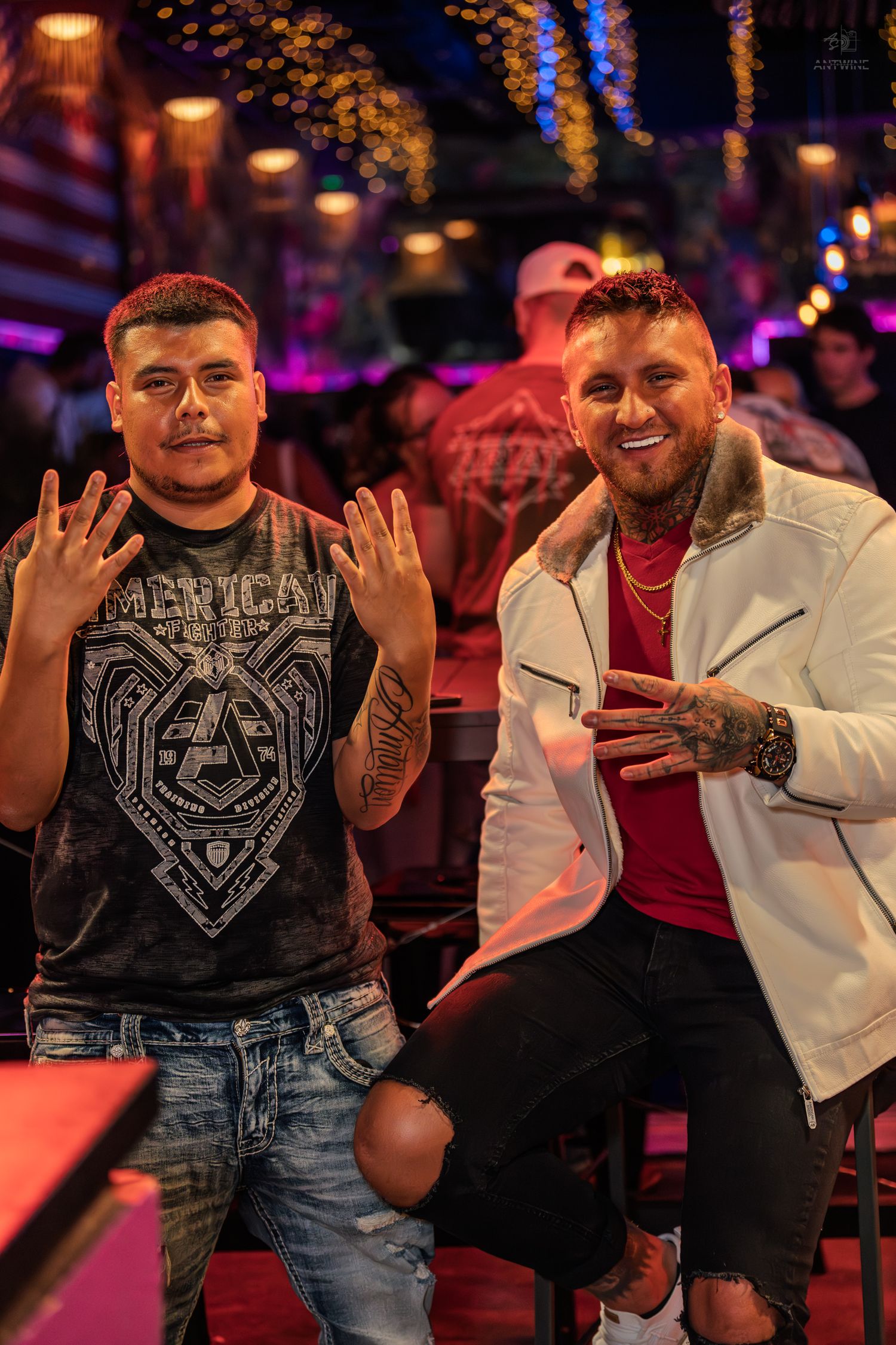 Two tattooed men posing and smiling together at a colorful and vibrant nightclub setting.