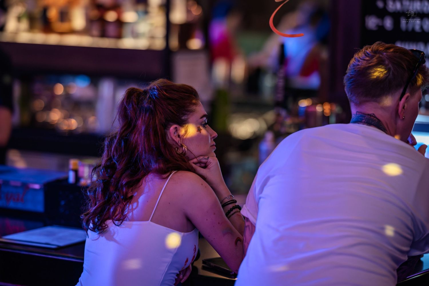 Young woman with ponytail sitting at a vibrant neon-lit bar conversing with a man.