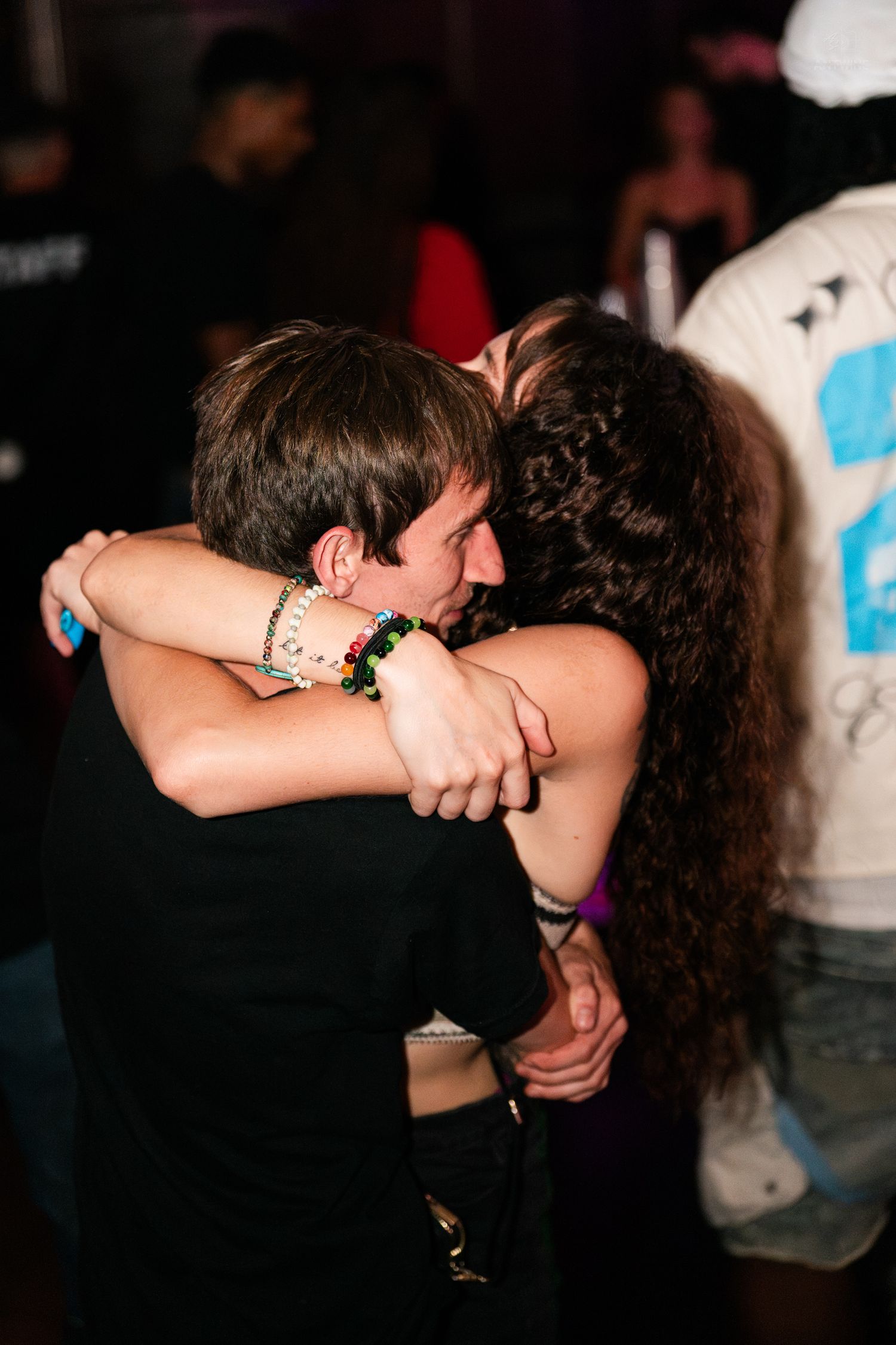 A couple shares a warm embrace on a crowded nightclub dance floor under dim lighting.