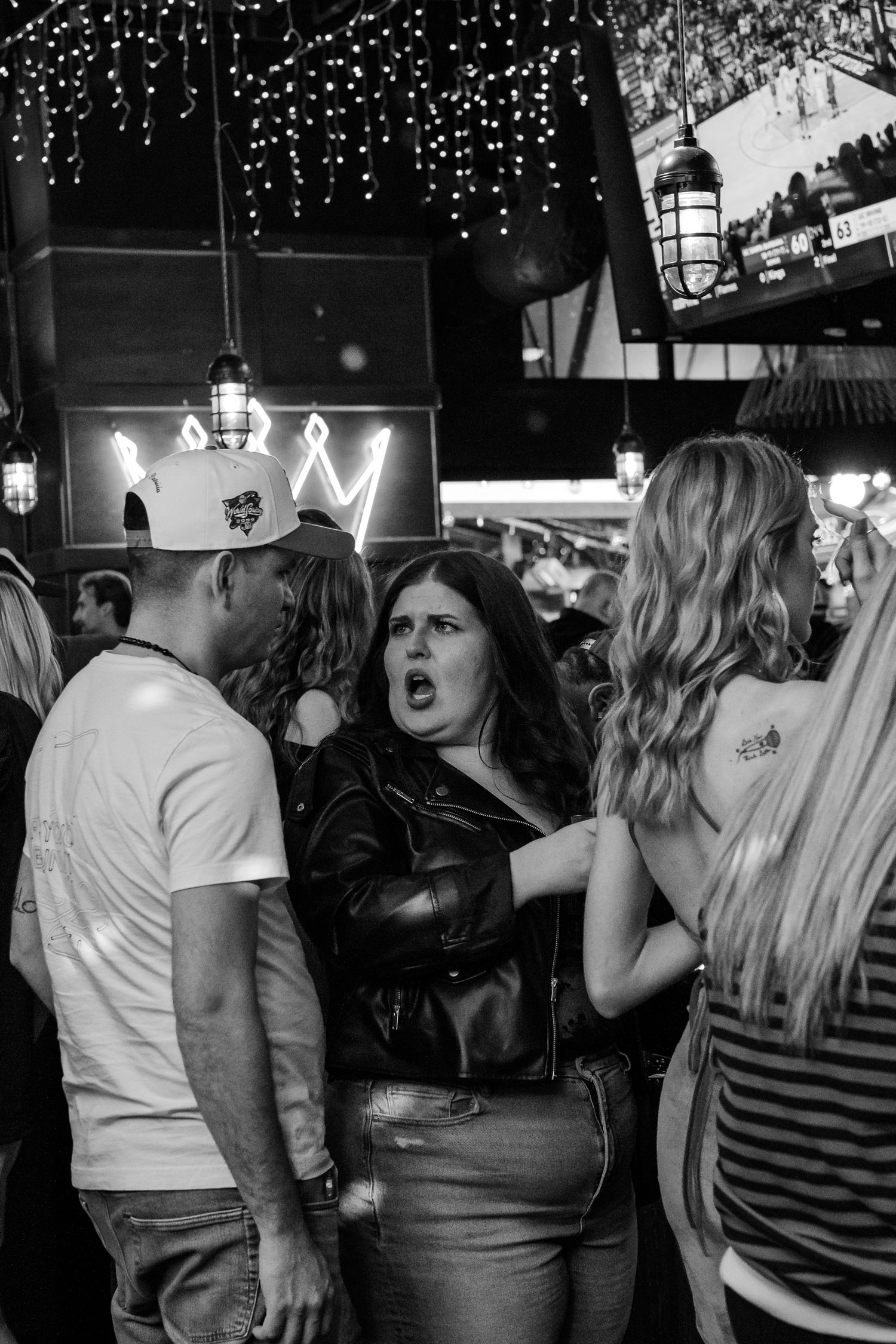 Black and white photo of a woman with an expressive face talking to people in a crowded nightclub.