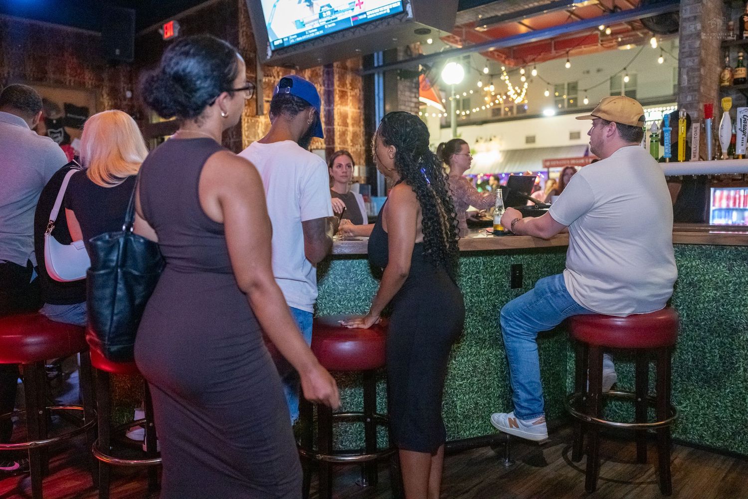 Diverse group of people socializing at a lively bar with red stools and green tiled counter.