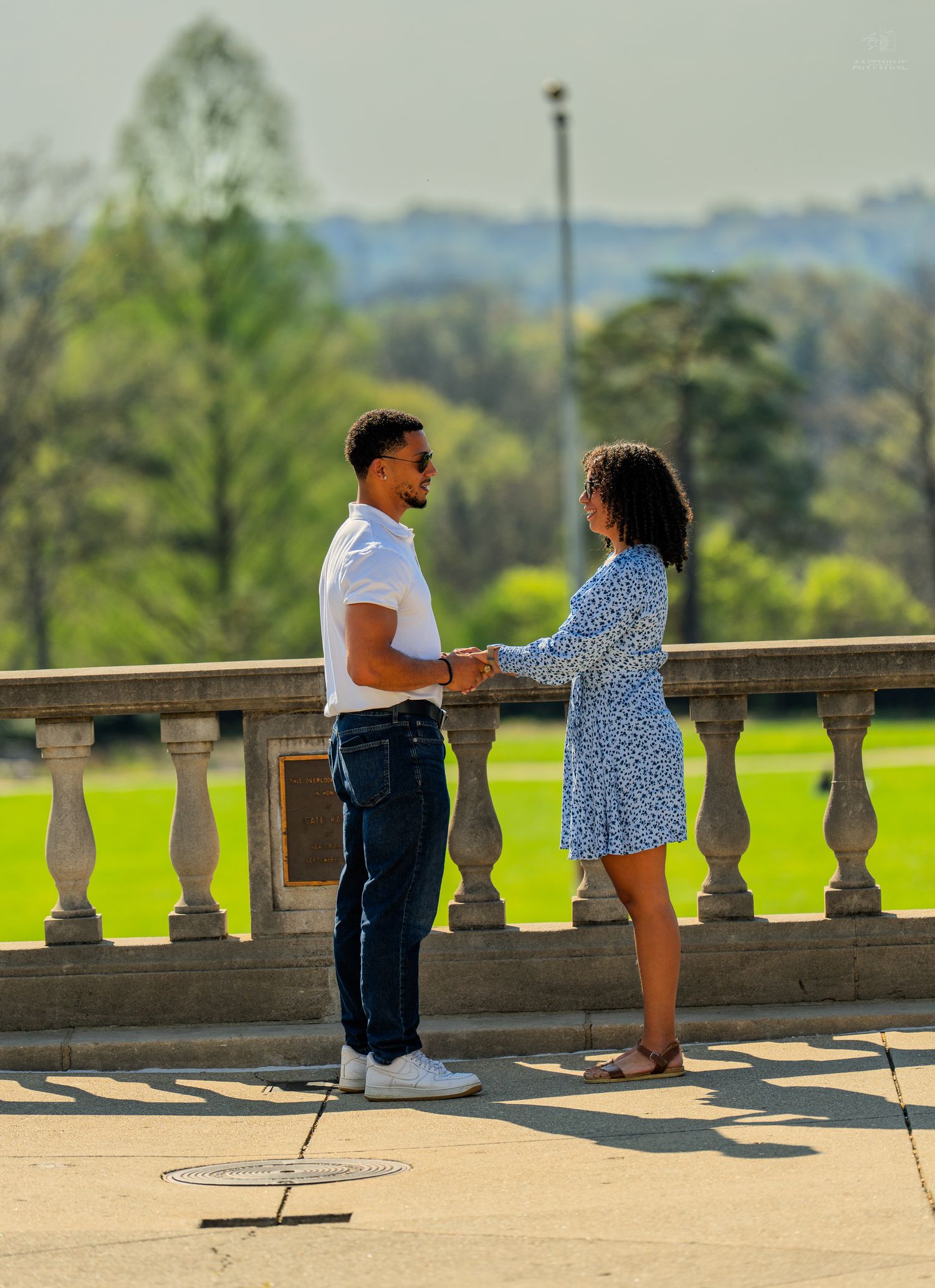 Couple holding hands on a warm sunny terrace with green lawn and trees in background.