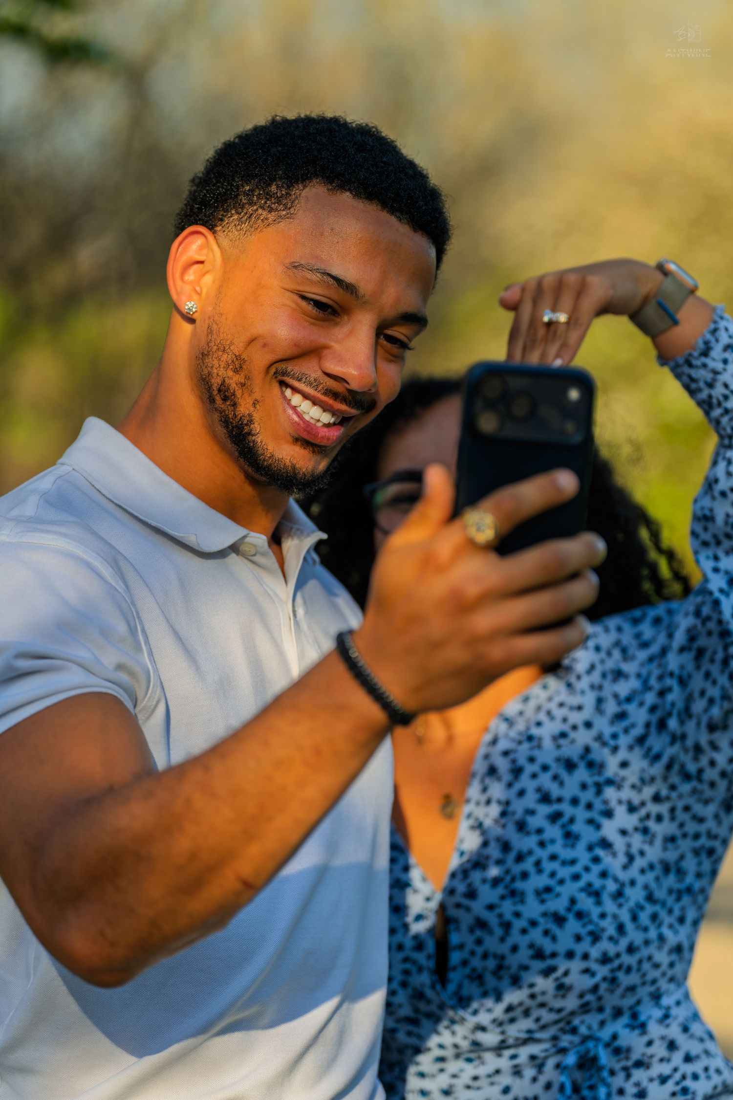 Smiling man taking a selfie with his partner in a sunny outdoor setting, woman resting head on his shoulder.