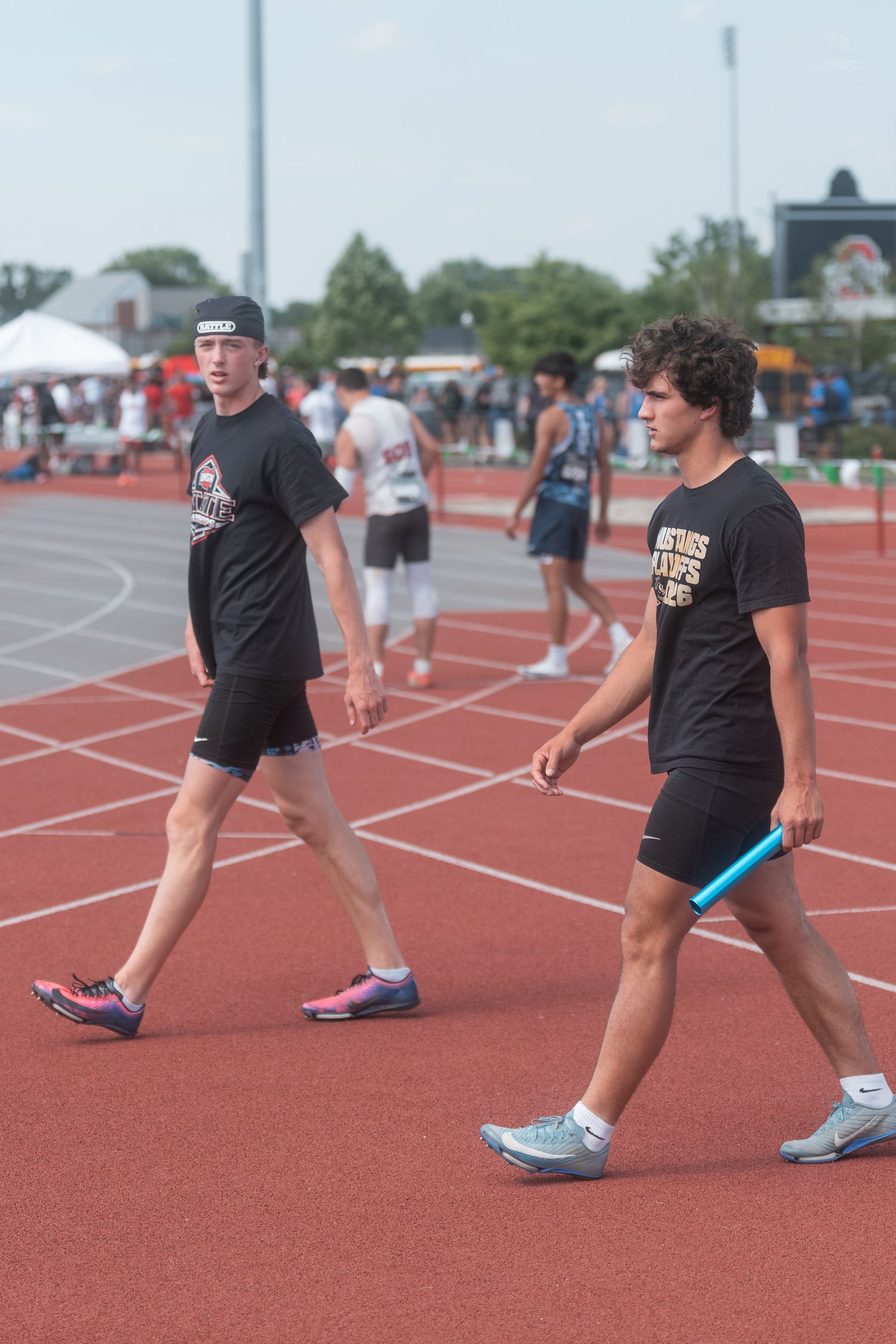 Two teenage male track athletes in black uniforms walk on a red track carrying a blue relay baton.