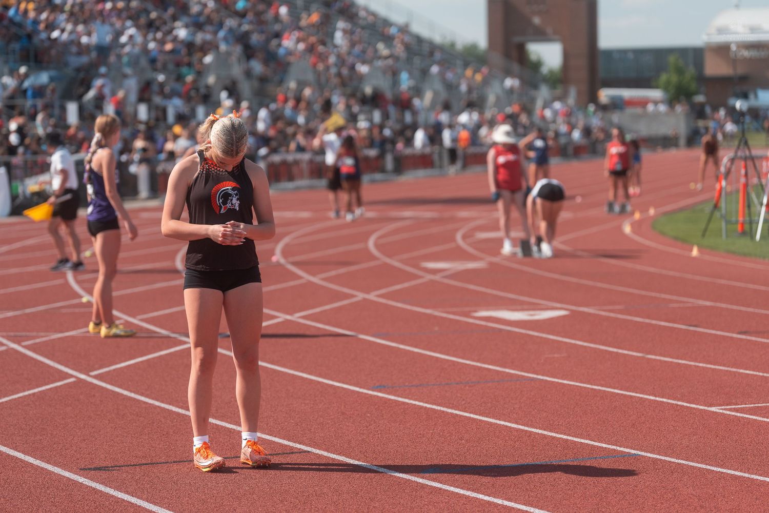 A female track athlete in an orange uniform checks her watch on an empty red track after completing a race.