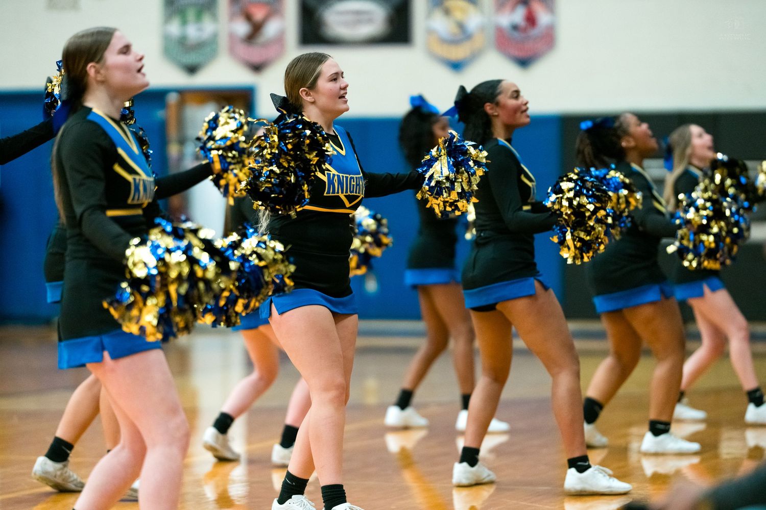 Smiling high school cheerleaders in black uniforms perform with gold and blue pom-poms at a basketball game.