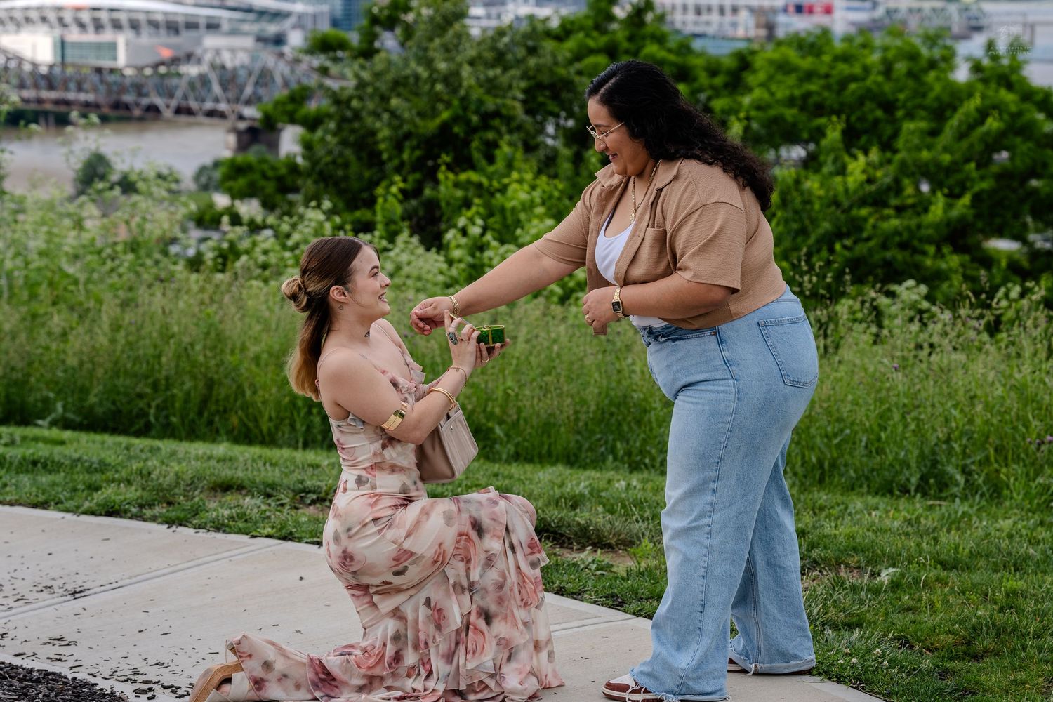 A woman kneels as another reaches toward her hand during an outdoor proposal in a garden setting.