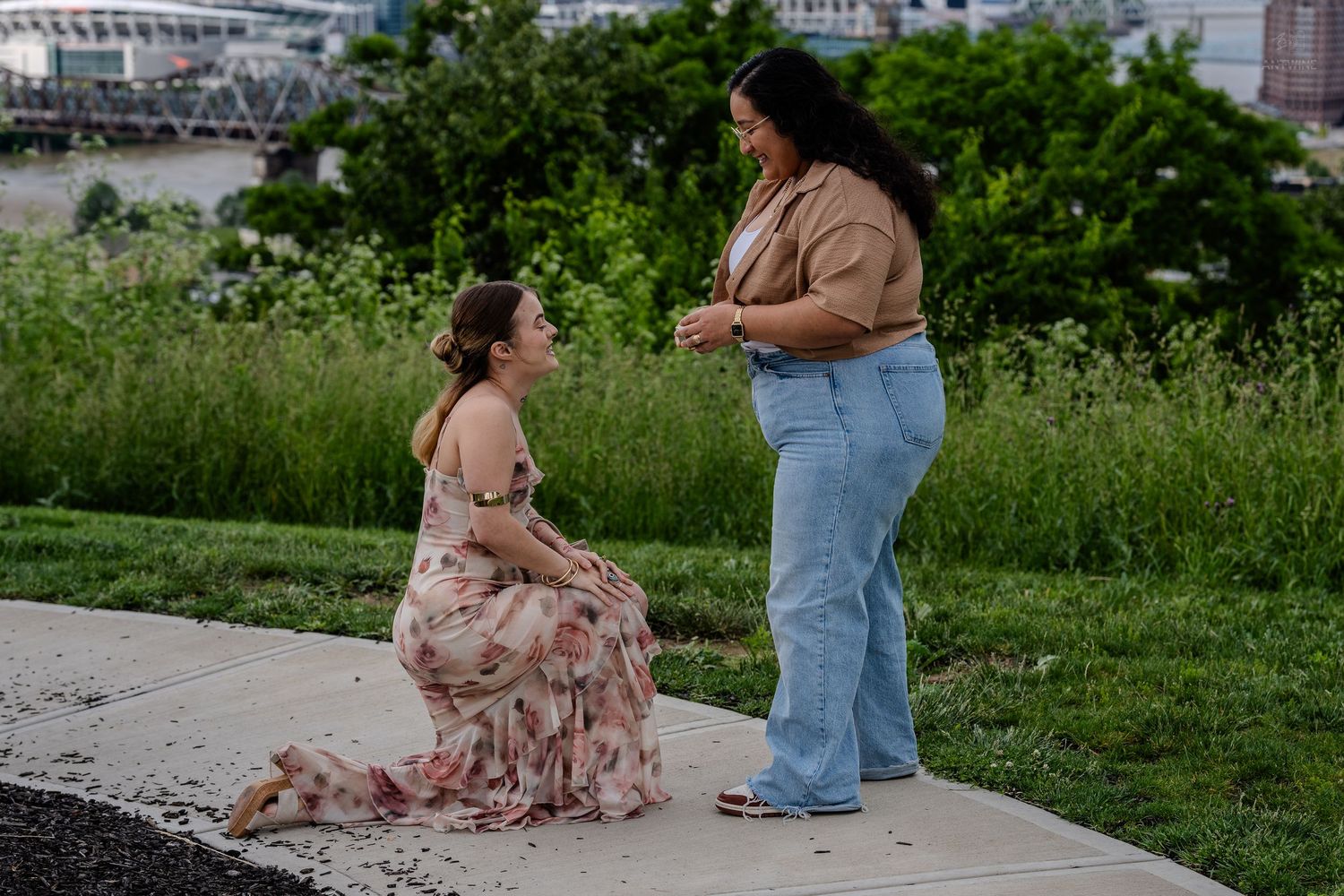 Woman in floral dress kneeling during outdoor marriage proposal with lush greenery and bridge behind.