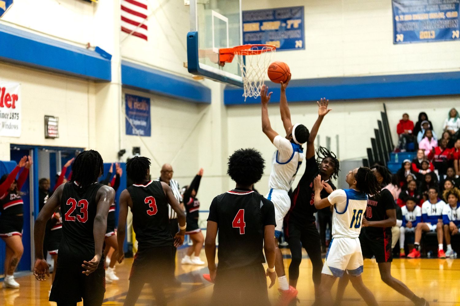 High school basketball players compete for a rebound during an intense game in a blue and white gymnasium.
