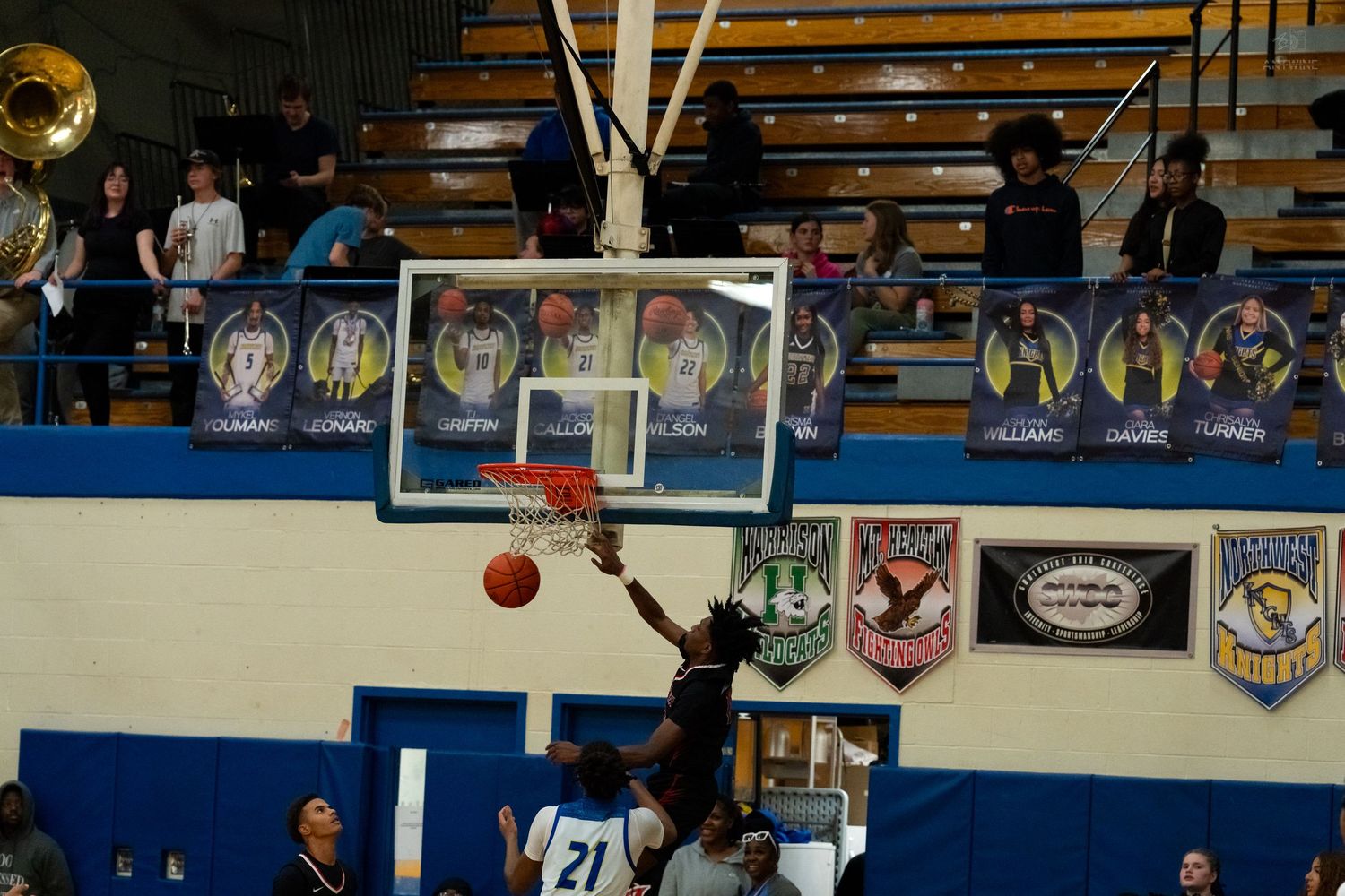 Basketball player #21 attempts a layup during a high school game in a blue and gold gymnasium.