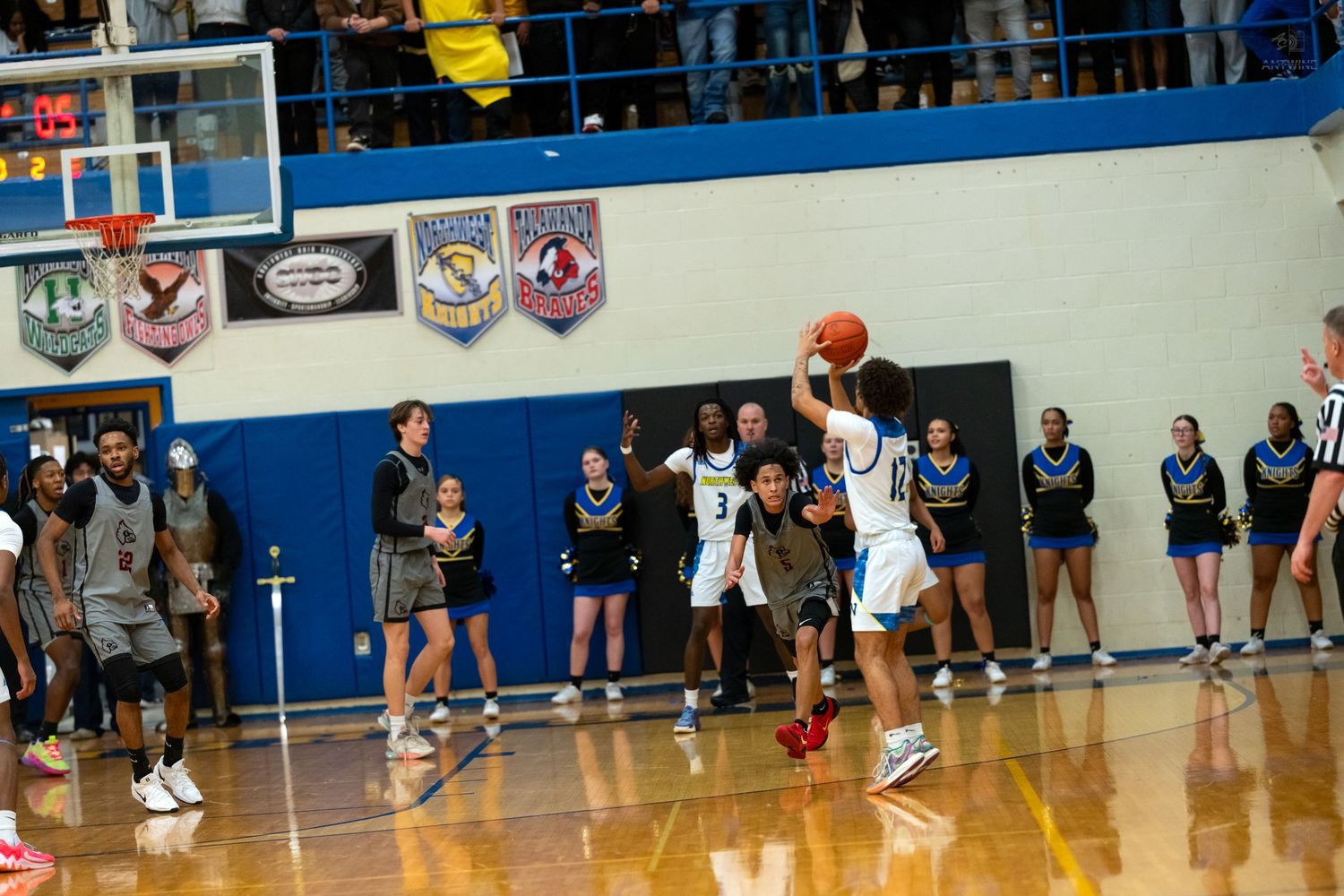 Basketball player shoots jump shot during high school game with team banners displayed on gymnasium wall.