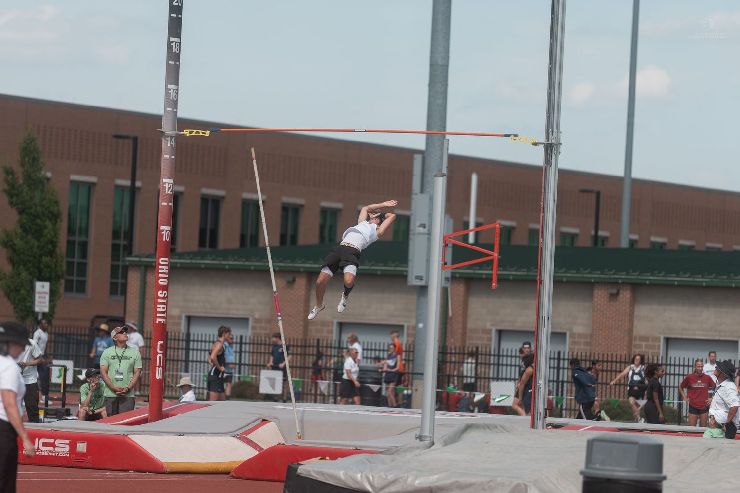 Pole vaulter in white uniform clears the bar at an outdoor track and field athletic competition.