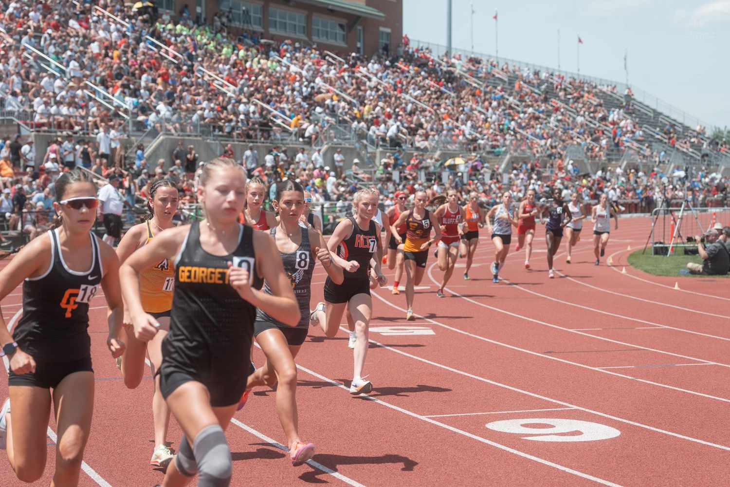 Female high school runners burst from the starting line during a track race at a large outdoor stadium meet.