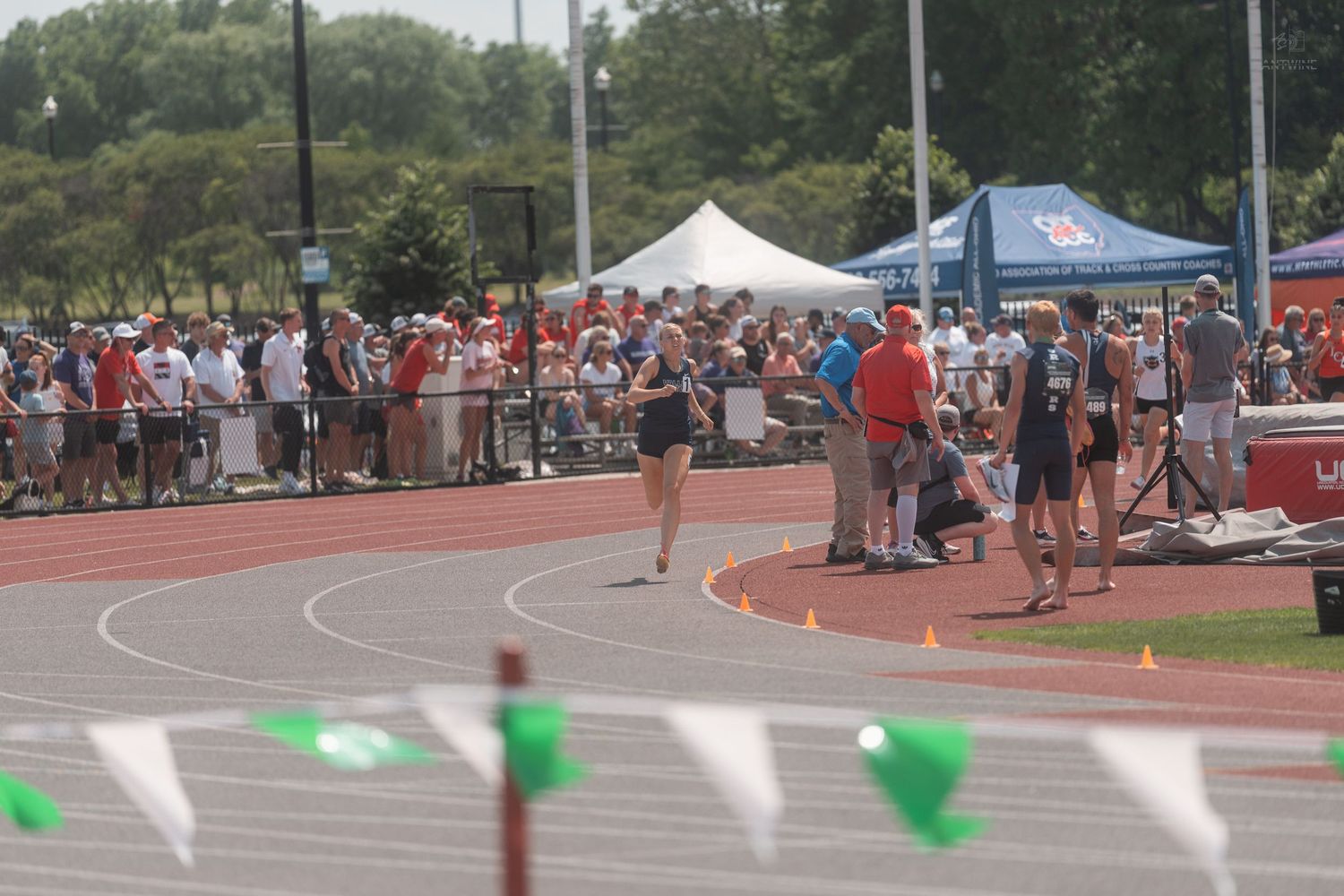 Track and field athletes running on oval track at crowded outdoor collegiate meet.