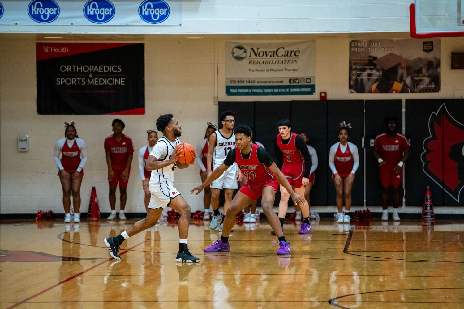 Basketball player dribbles upcourt during a high school game with cheerleaders and sponsors visible in background.