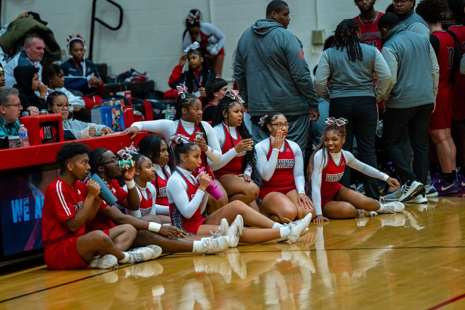 Cheerleaders in red and white uniforms sit along the sideline of an indoor basketball court.