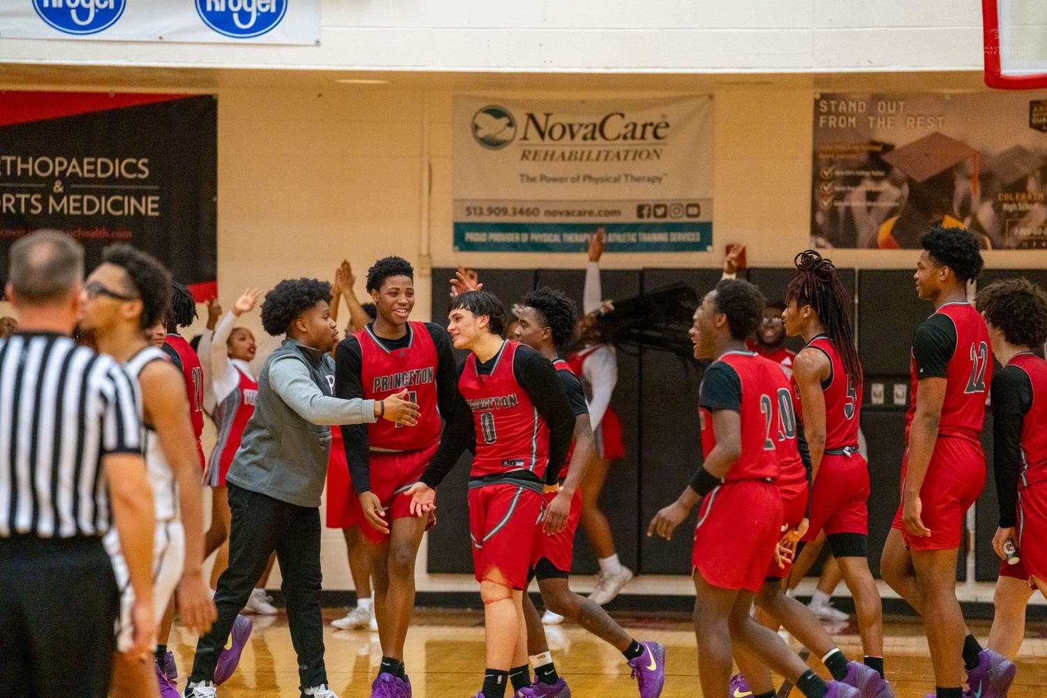 Red-uniformed basketball team celebrates together on court near NovaCare Rehabilitation banner during high school game.