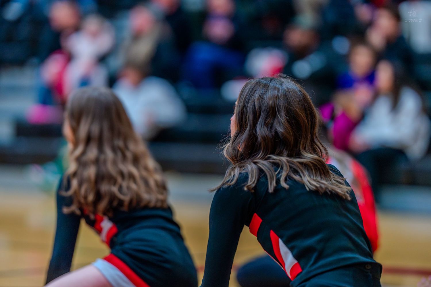 Two cheerleaders in black and red uniforms sit courtside during a high school basketball game.