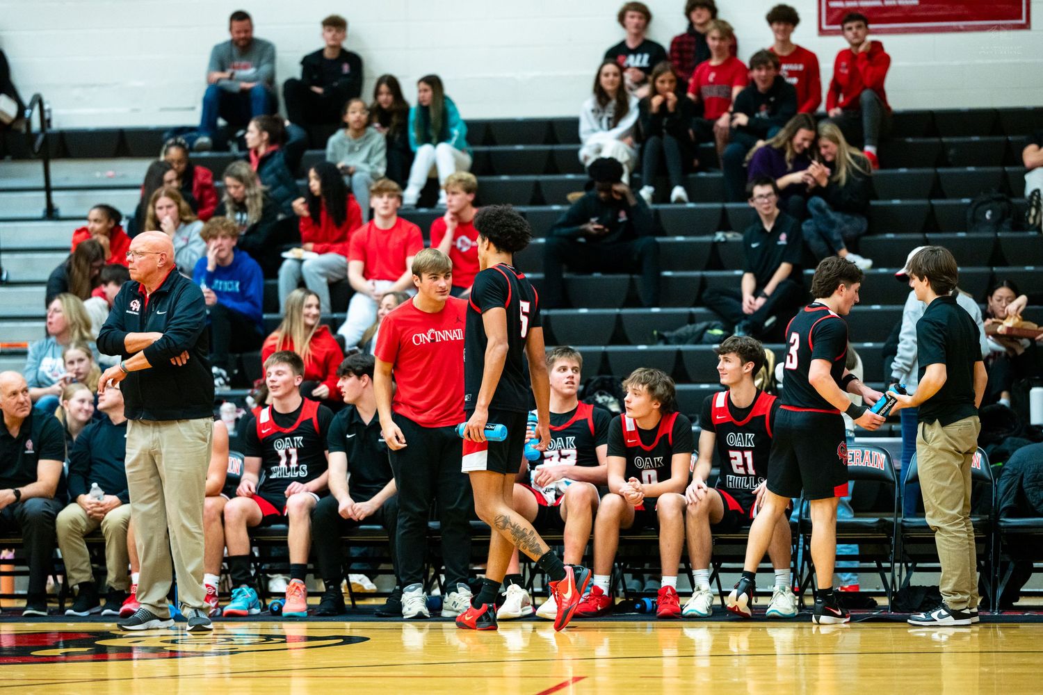 High school basketball team huddles on sideline bench during timeout in red uniforms.