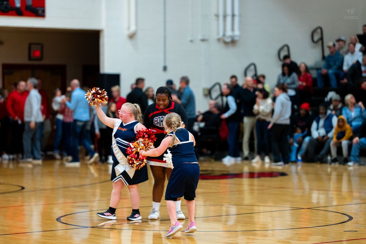 Young cheerleaders in navy uniforms practice routines on basketball court with crowd watching in background.