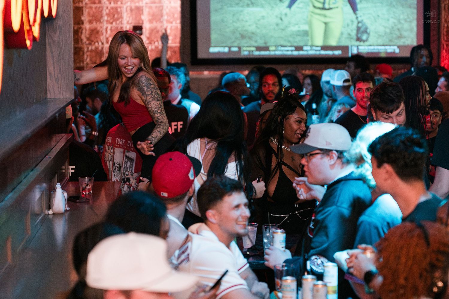 Lively sports bar scene with patrons socializing under a large projection screen.