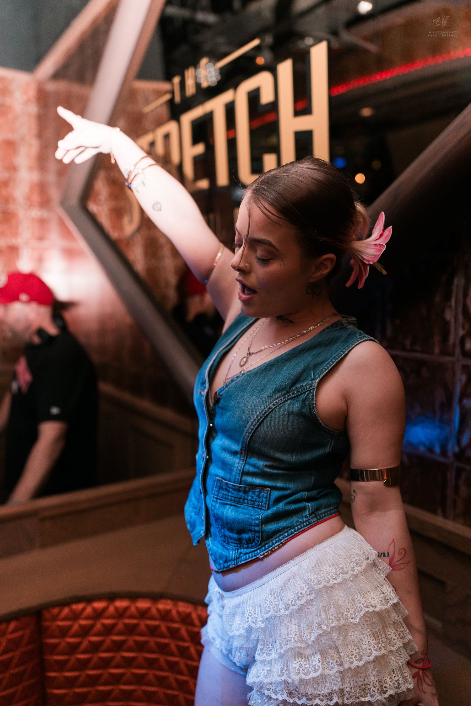 Young woman in denim vest and white ruffled shorts dancing with arm raised at a trendy bar venue.