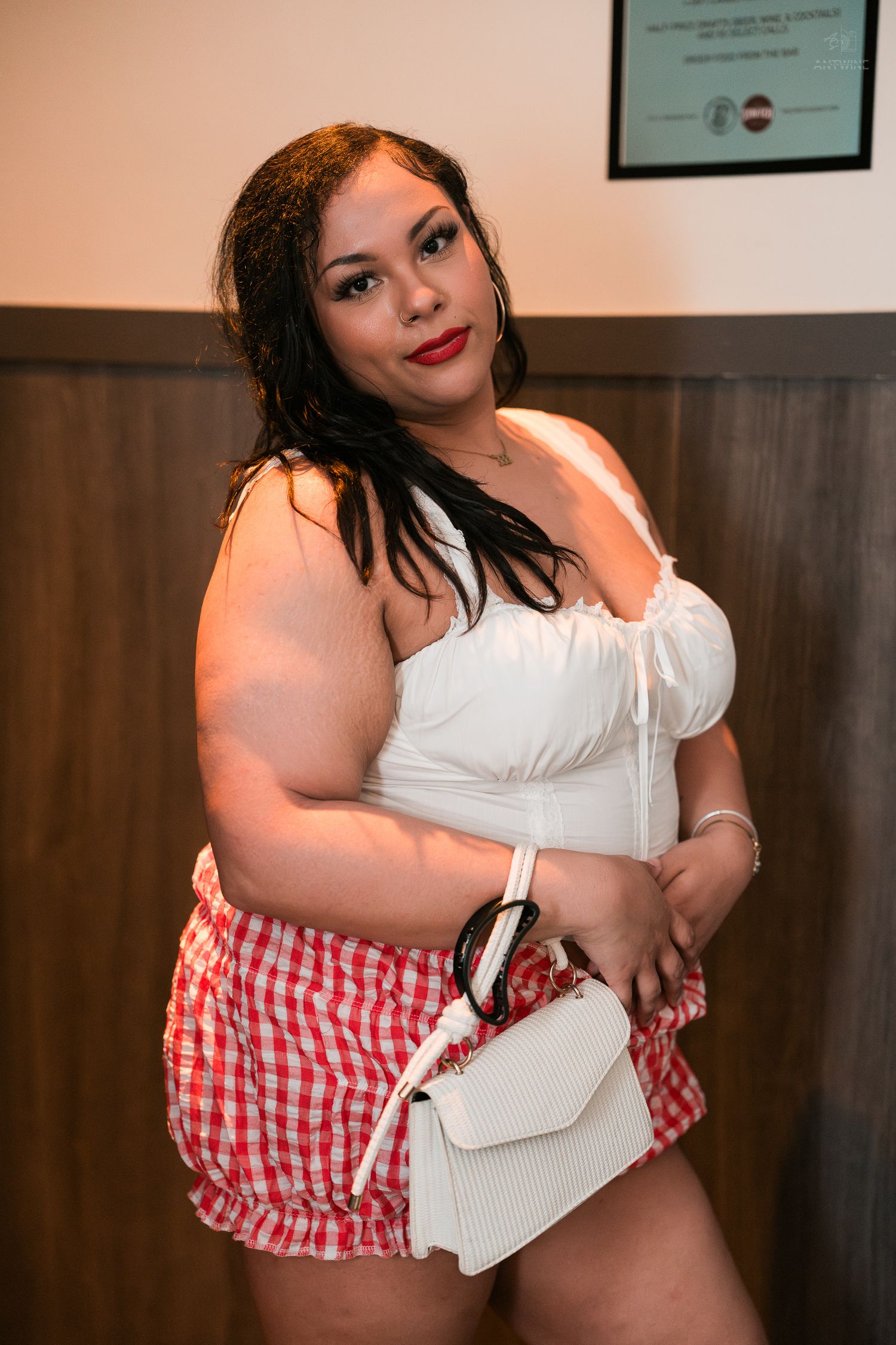 Plus-size woman in white crop top and red gingham skirt posing with white handbag.