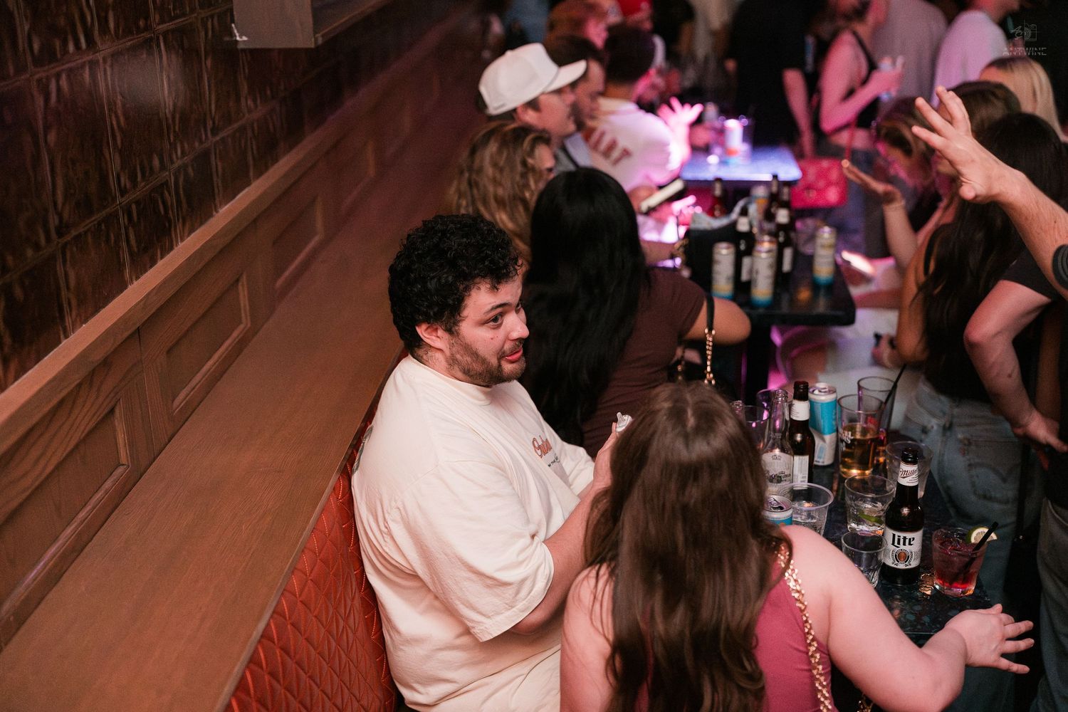 Young man in white shirt chatting with friends at a busy bar counter with drinks.