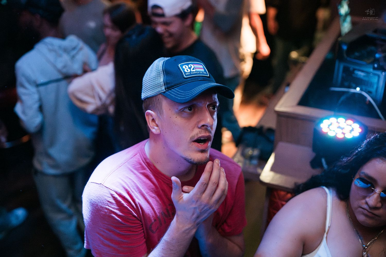 Man in blue trucker hat looks surprised at a lively nightclub event with stage lighting behind him.