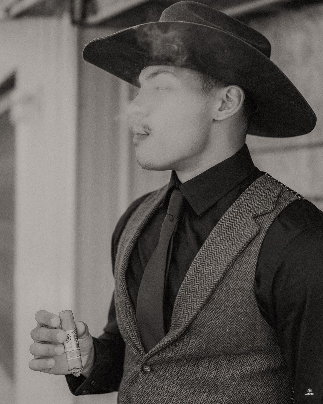 Black and white portrait of a groom in a cowboy hat and vest smoking a cigar at a wedding.