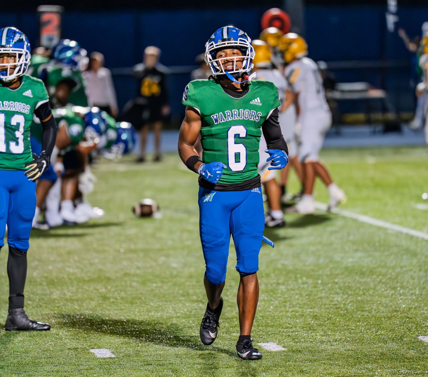 High school football player number 6 in green Warriors jersey jogs on field during a night game.