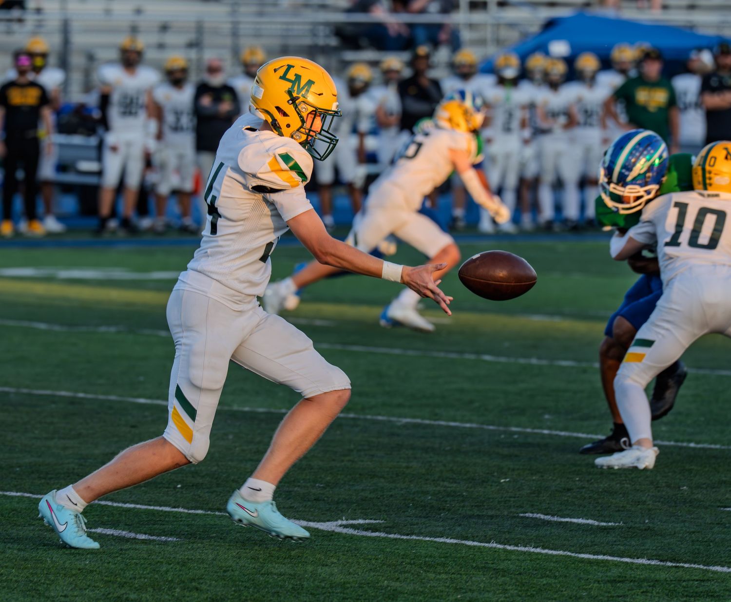 A yellow and white uniformed player punts the football during a high school football game on blue turf.