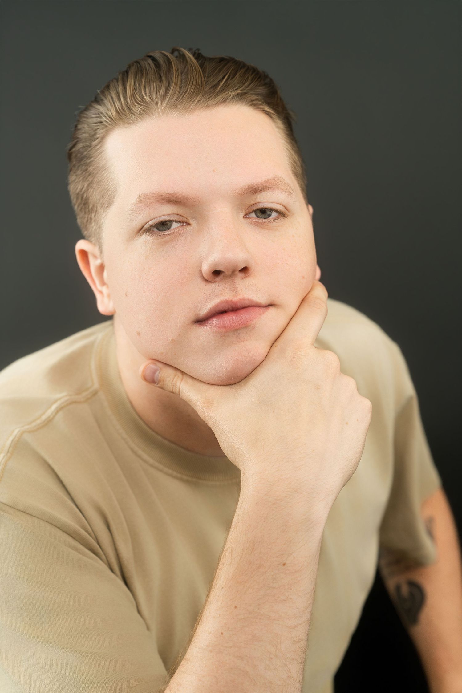Young man in beige t-shirt resting chin on hand in thoughtful pose against dark gray background.