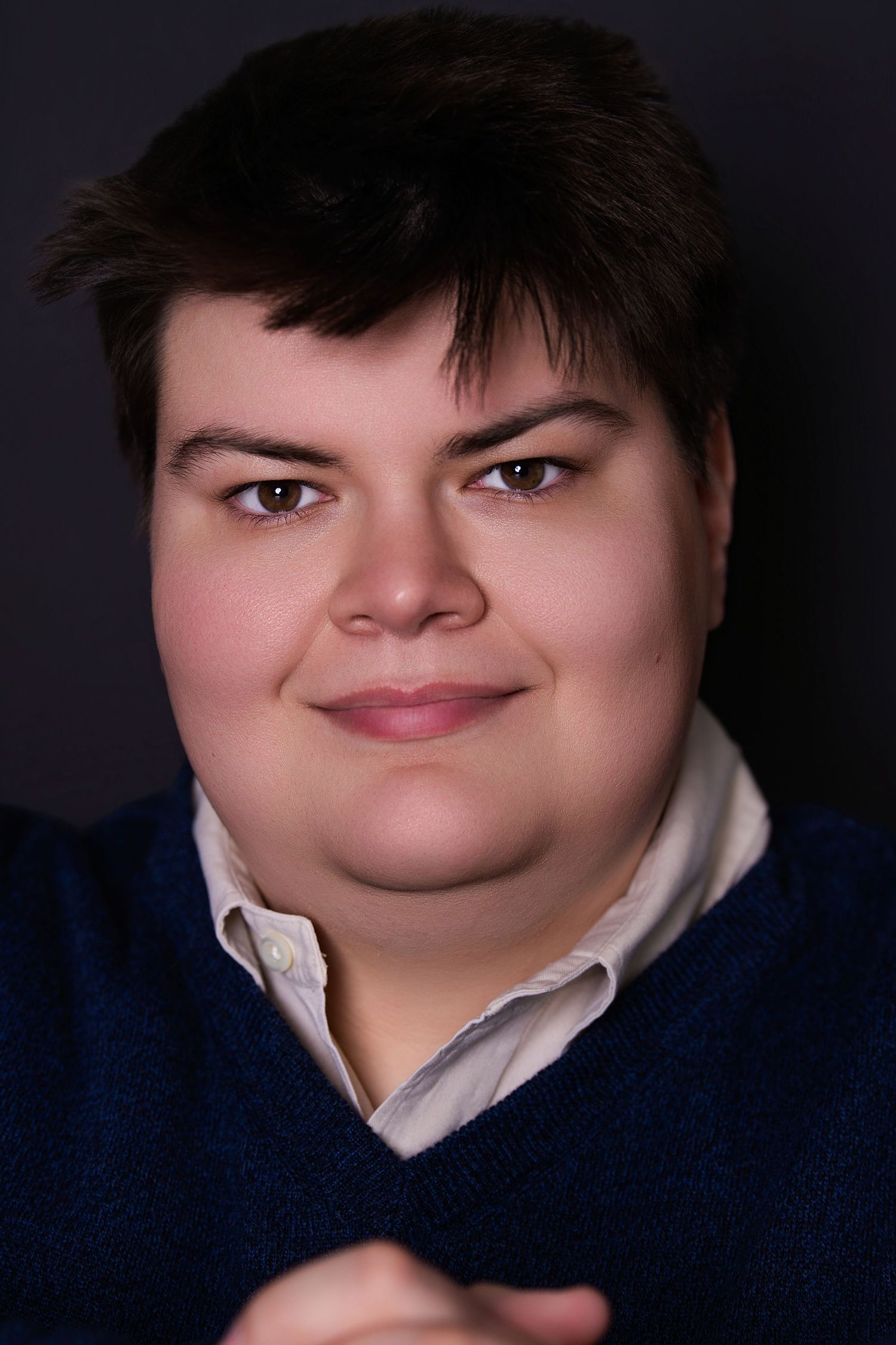 Young person with short dark hair in navy sweater smiles warmly for a studio portrait.