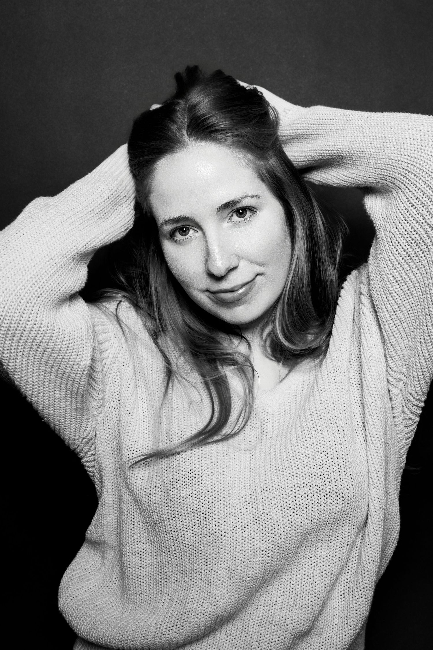 Black and white studio portrait of a young woman posing with hands in hair wearing a knit sweater.