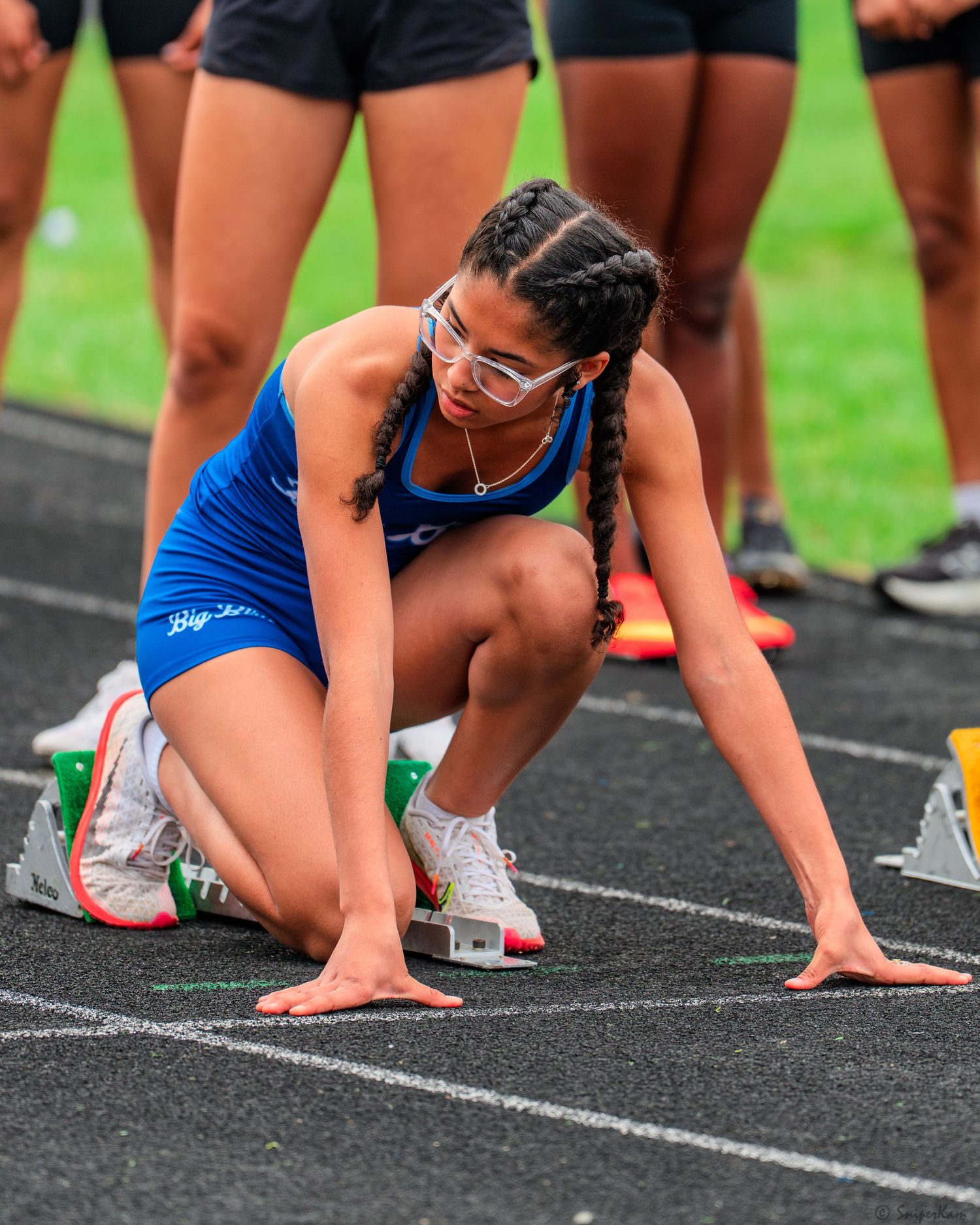 Female athlete in blue uniform with braids poised in starting blocks on a track field.