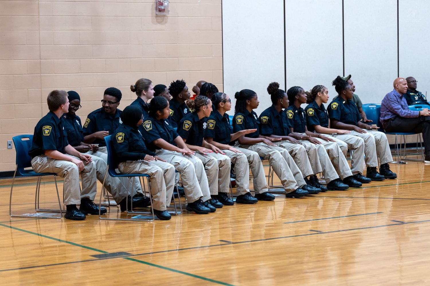 Group of uniformed officers seated in rows on gym floor during an official ceremony or graduation event.