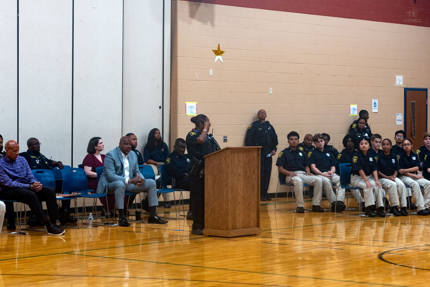 Students and officials gather in a school gymnasium for a formal ceremony with officers seated along the walls.