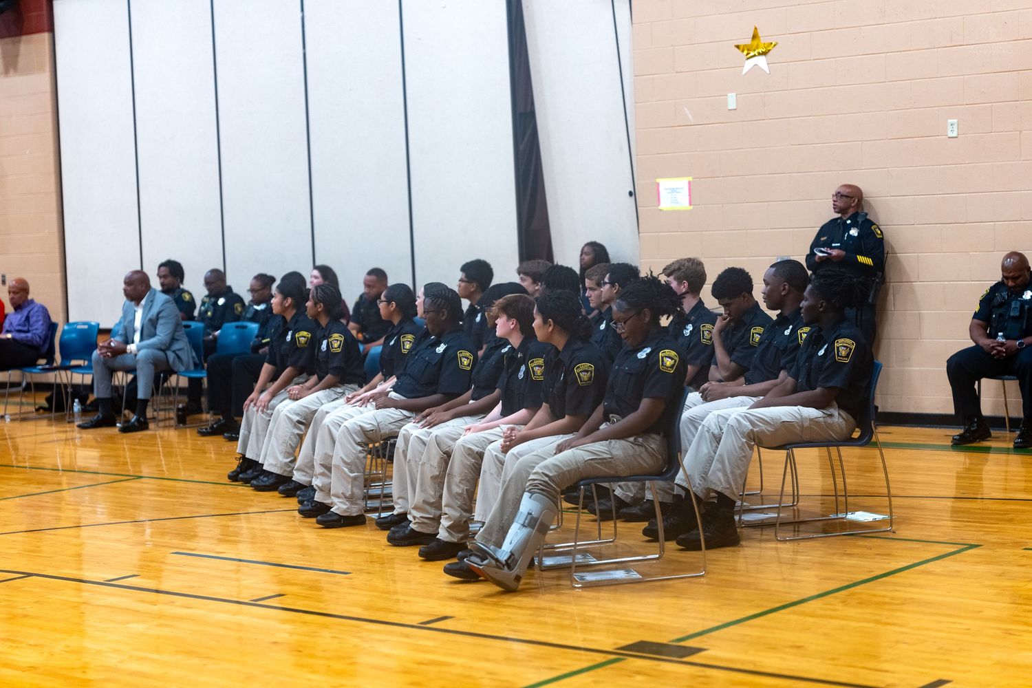 Group of youth cadets in uniforms seated in gymnasium chairs during a formal ceremony or graduation event.