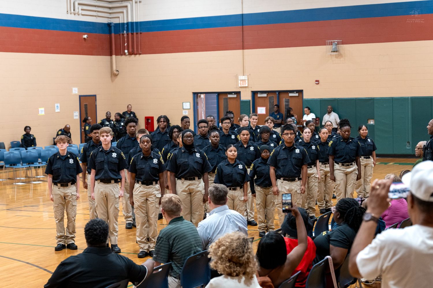 Group of students in matching black polo shirts and khaki pants standing in formation in a school gymnasium.