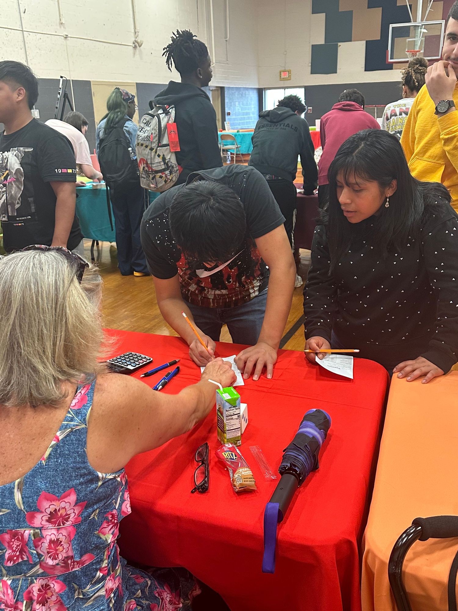 Students and adults interact at a colorful school fair event, gathered around a red table in a gymnasium.