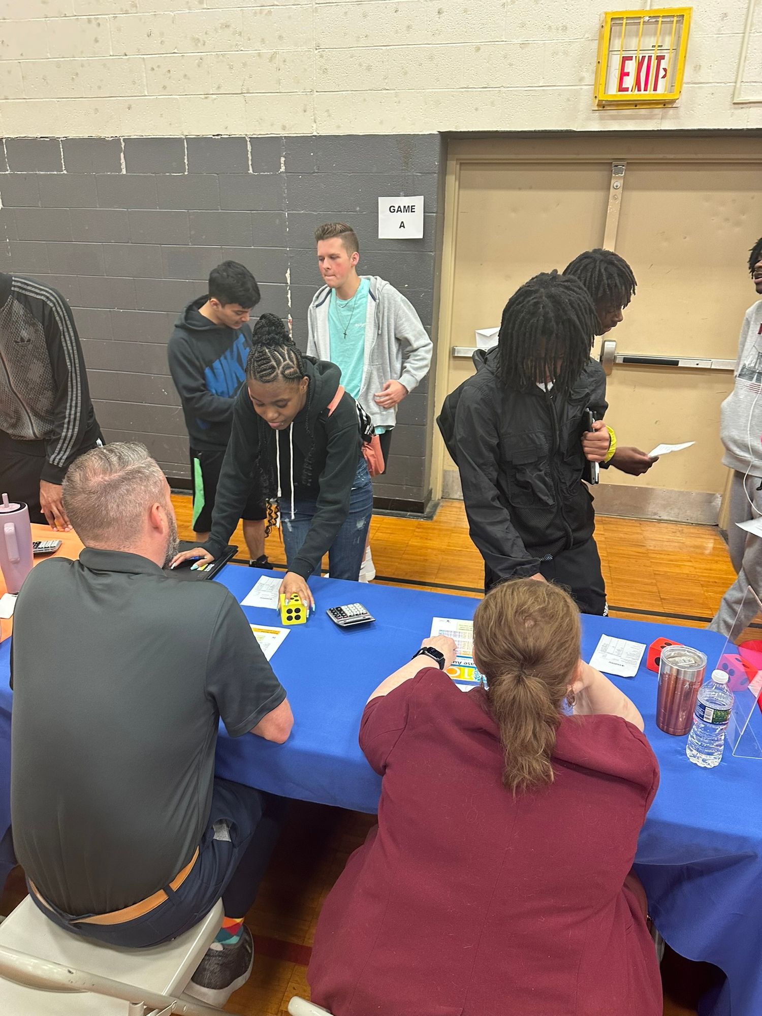 Students gather around a blue table at an indoor school career fair event in a gymnasium.
