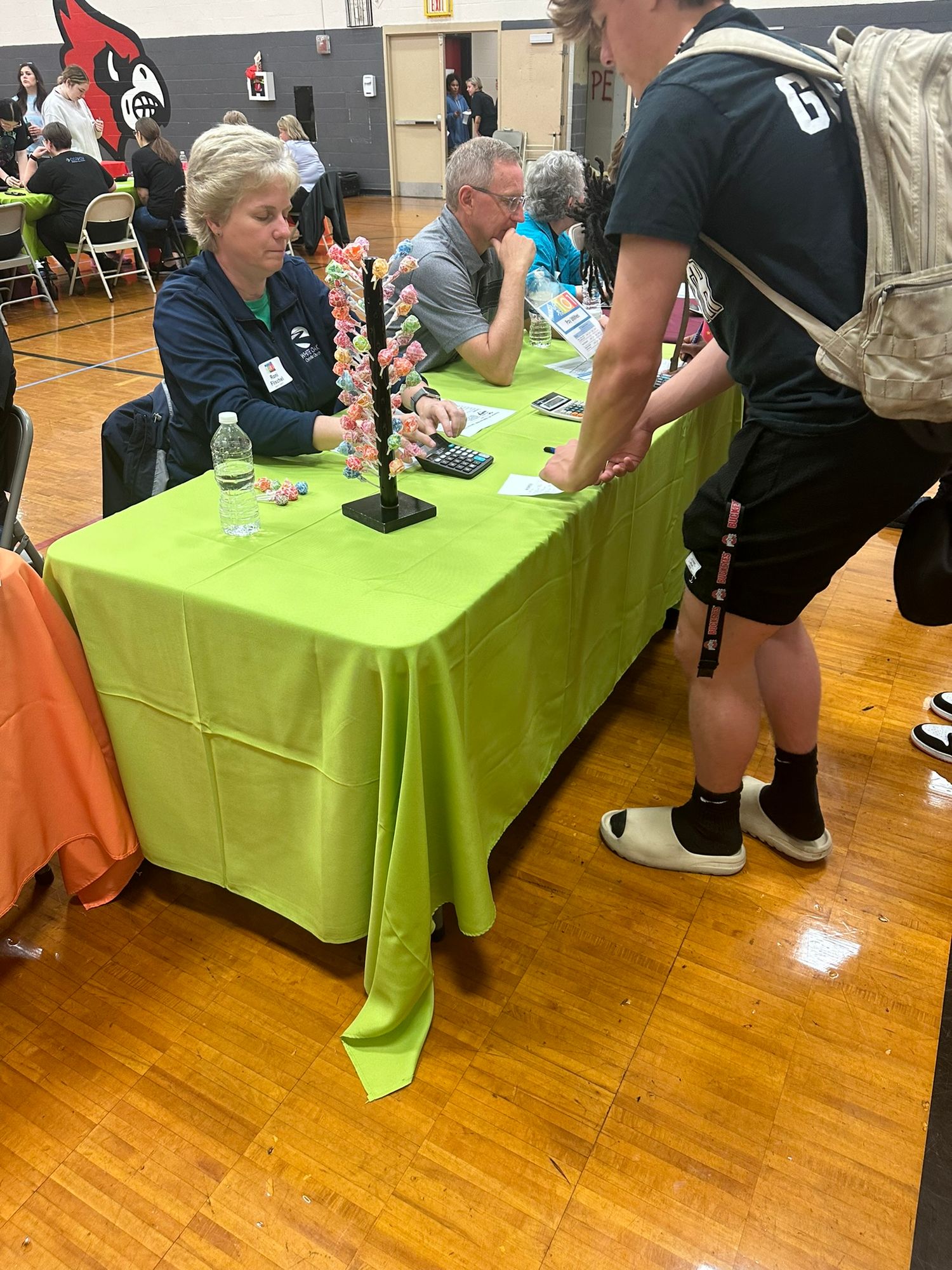 People gathered around a green tablecloth booth at an indoor school gymnasium career or health fair event.