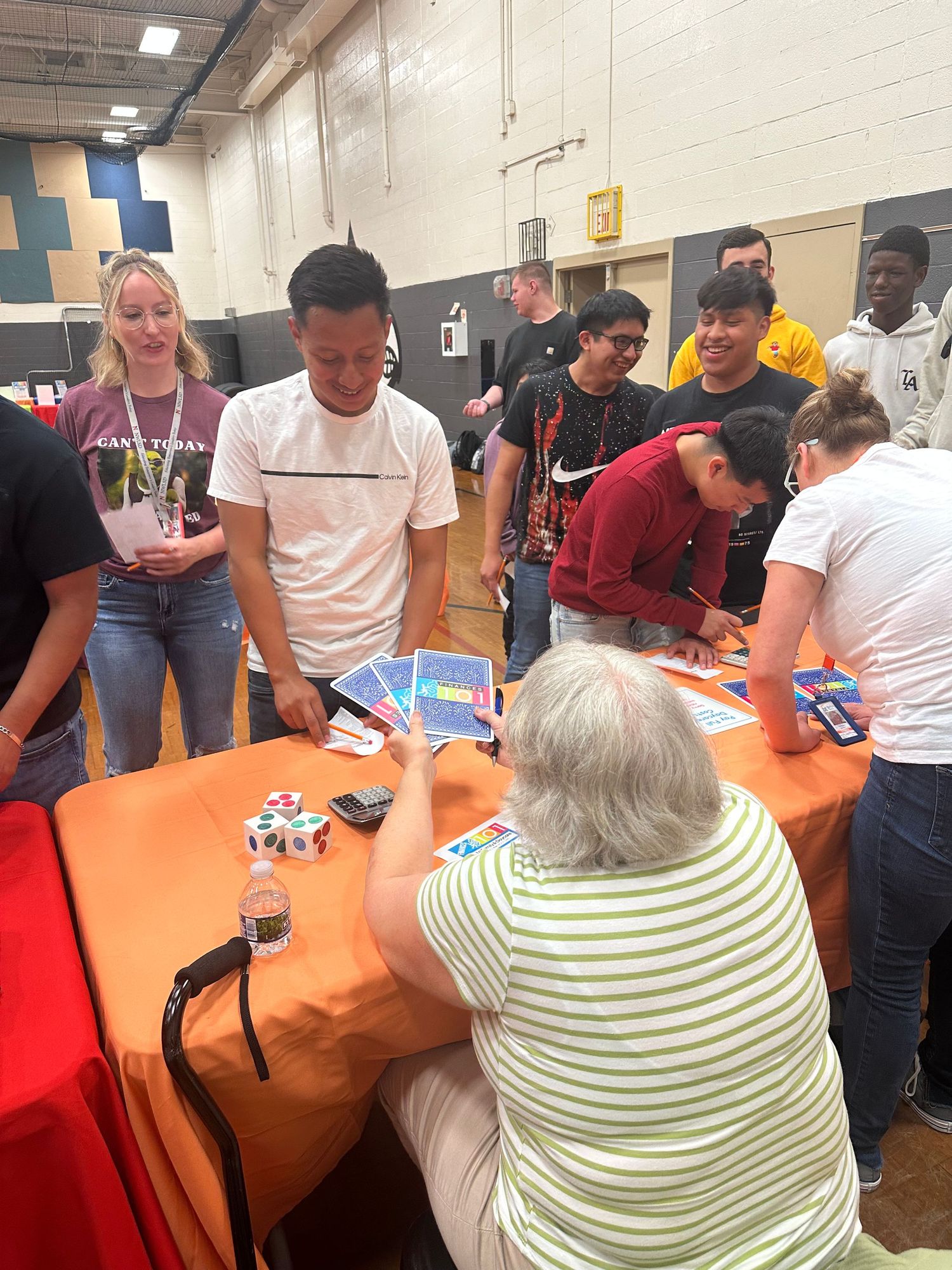 Students gather around an orange table in a school gym, engaging with an elderly woman during an educational fair event.