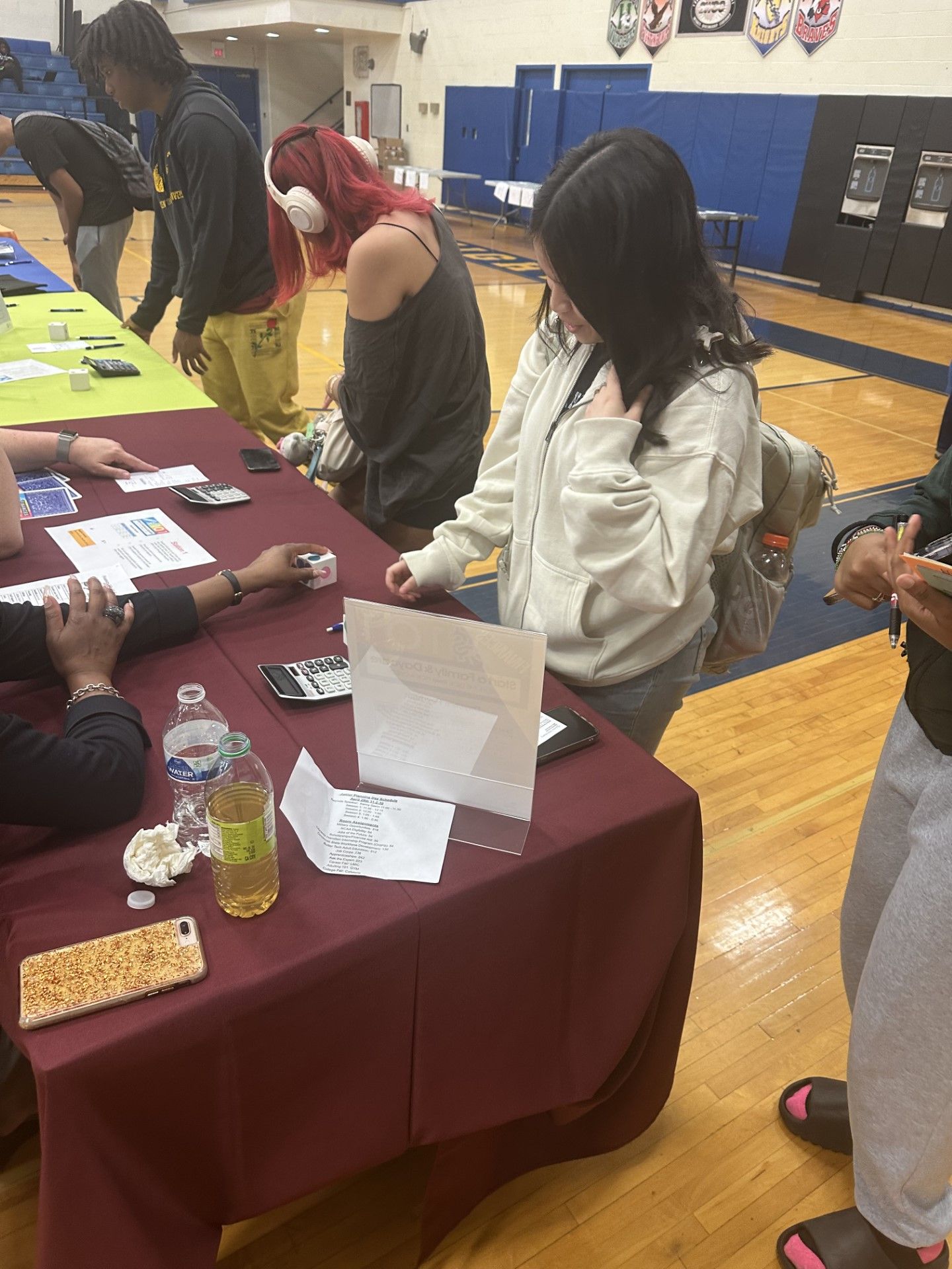 Students gather at a maroon-draped table in a school gymnasium during a college or career fair event.