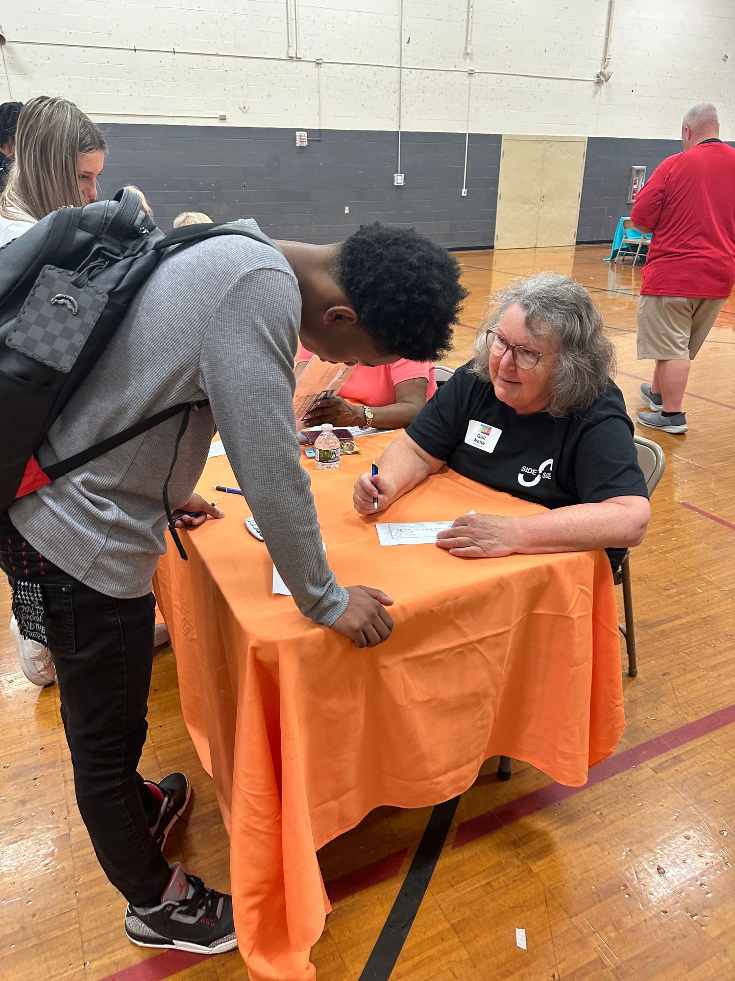 Older woman with glasses assists a student signing paperwork at an orange-covered table inside a school gymnasium event.