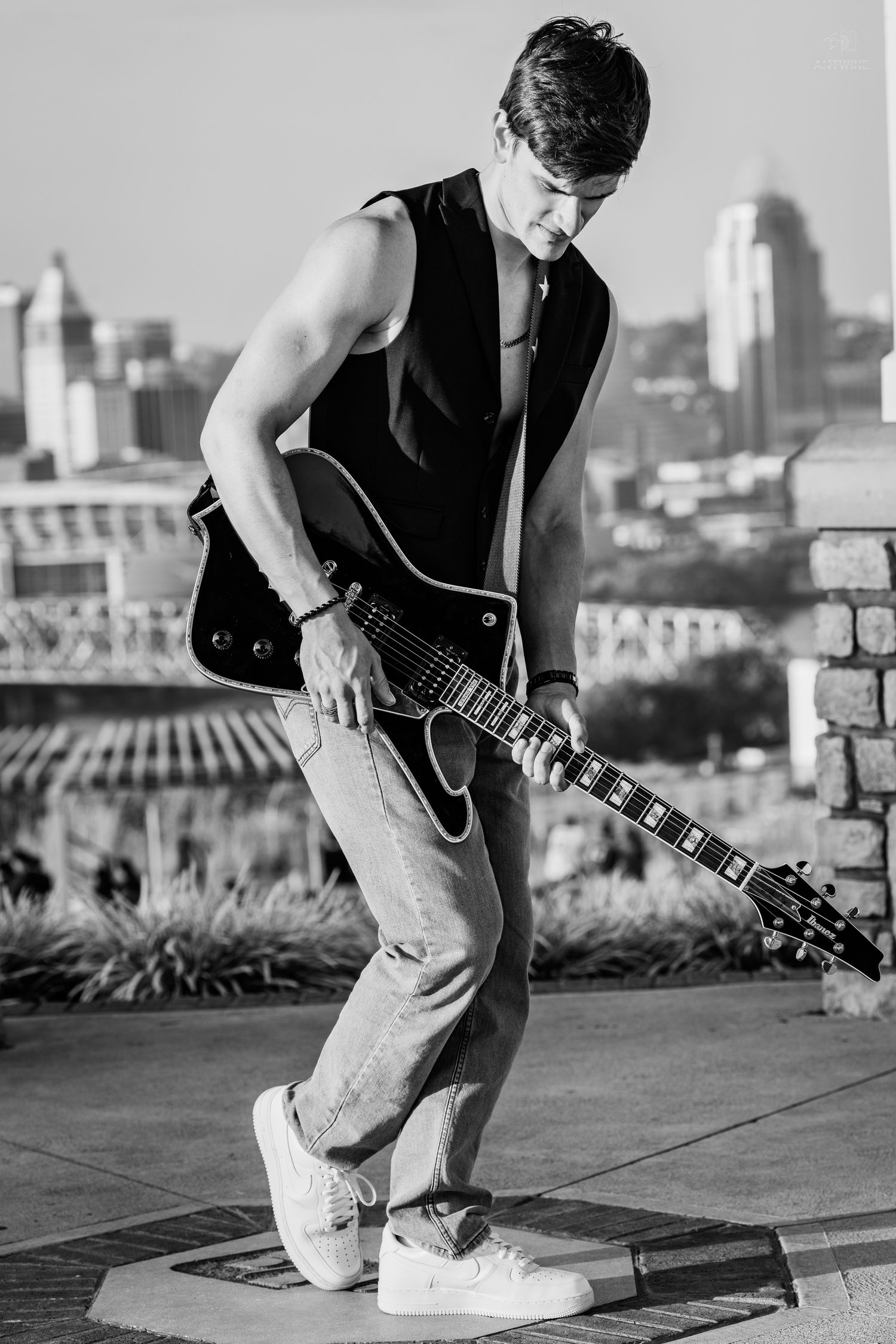 Young man playing electric guitar outdoors in black and white with city skyline background.
